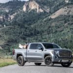 A light gray 2021 GMC Sierra Denali has a dirt bike in the bed while parked in front of tree-covered mountains.