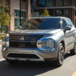 A silver 2022 Mitsubishi Outlander is driving on a two way road through an urban area.