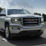 A low angle shows a popular used GMC truck, a white 2016 GMC Sierra 1500 SLT, driving in front of a field and trees.