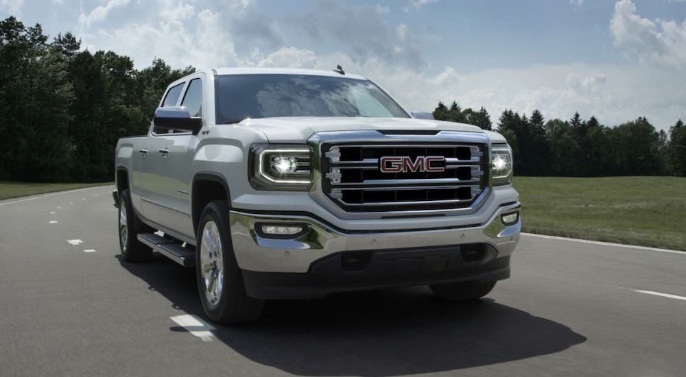 A low angle shows a popular used GMC truck, a white 2016 GMC Sierra 1500 SLT, driving in front of a field and trees.