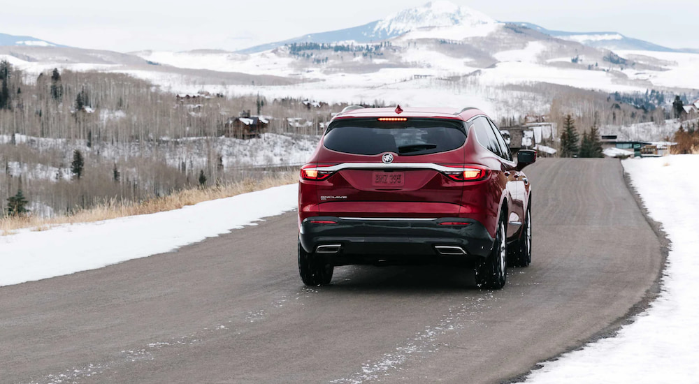 A red 2021 Buick Enclave is shown from the back driving on an open road in front of snowy mountains.