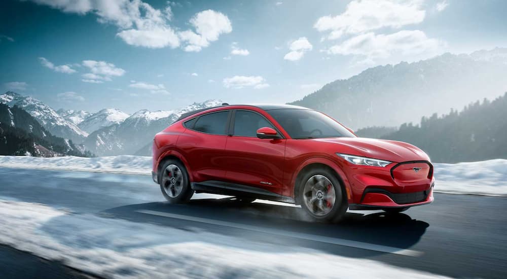 A red 2021 Ford Mustang Mach-E is driving on a snow covered road past a mountain.