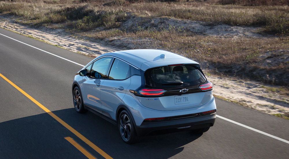 A light blue Chevy Bolt EV is driving down an open road next to a sand dune.