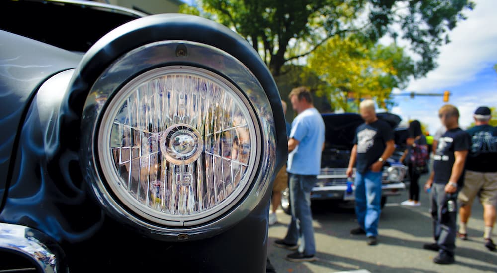 A close up shows a headlight and people at a car meet.