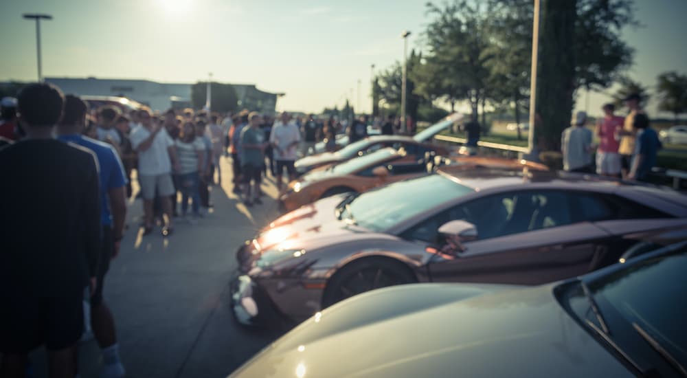 A row of cars and people are shown on a sunny day at a car meet.