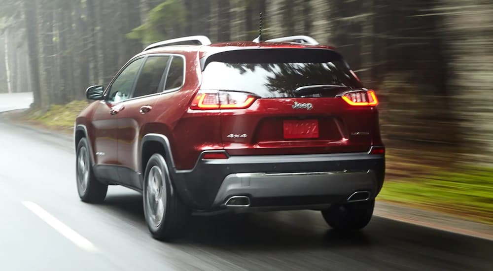 A red 2021 Jeep Cherokee is shown from the rear driving on a road next a forest.
