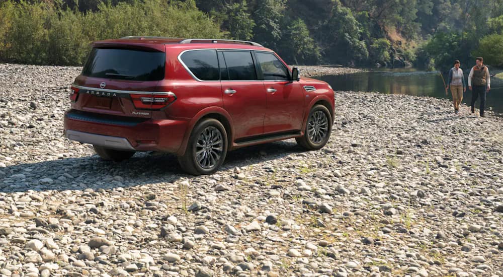 A red 2021 Nissan Armada is parked on stones next to a river as a couple walks along the bank.