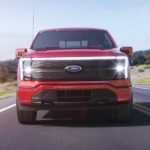 A red 2022 Ford F-150 Lightning Lariat is shown from the front driving down a road.