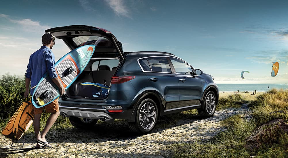 A blue 2021 Kia Sportage is shown parked at the beach as a surfer takes items from the open lift gate.