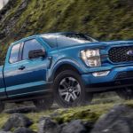A blue 2021 Ford F-150 STX is shown driving past a rock face.