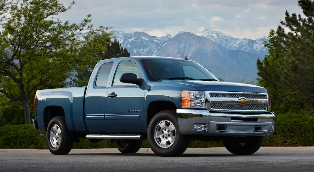 A blue 2012 Chevy Silverado is parked on pavement in front of mountains.