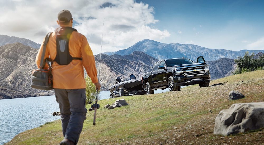 A black 2018 Chevy Silverado 1500 High Country with a boat on a trailer is parked next to a man with a fishing pole.