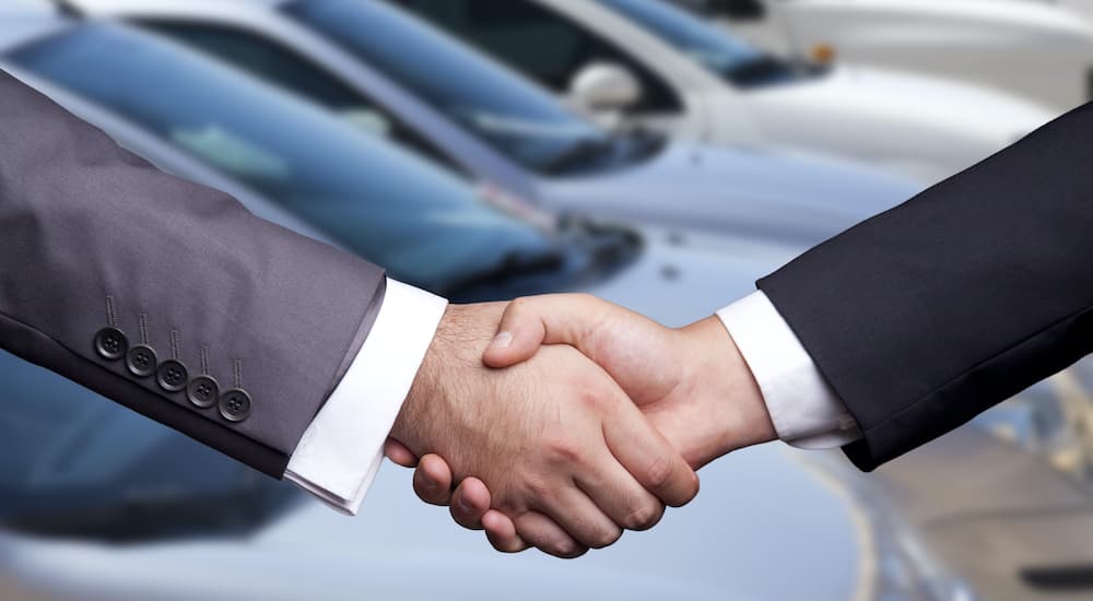 A salesman is shaking hands with a car buyer at a used car lot.