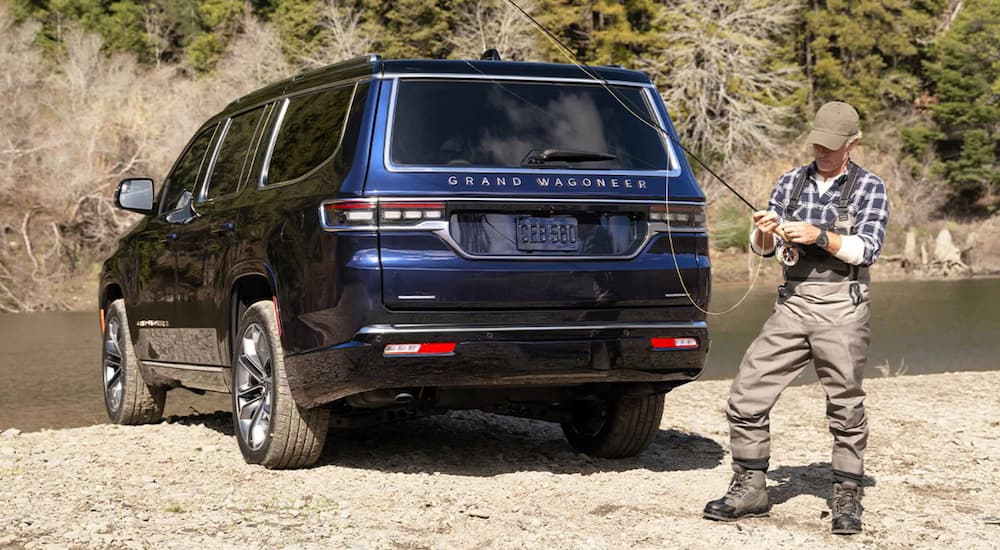 A blue 2021 Grand Wagoneer is shown from the rear parked on a river bank next to a fisherman.