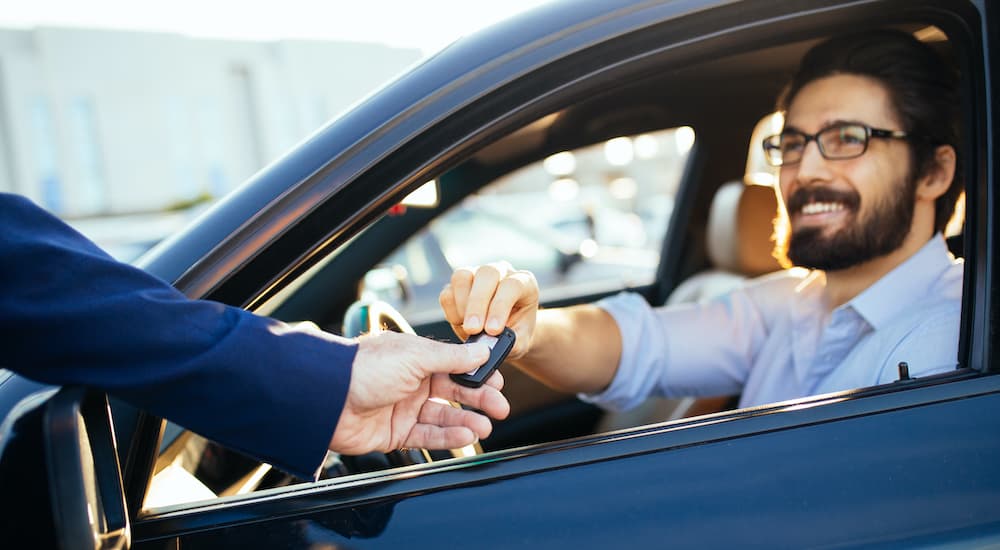 A man is sitting in the drivers seat of a car as a salesman passes him a key.