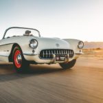 A white 1957 Chevy Corvette is shown driving down a sunny road.