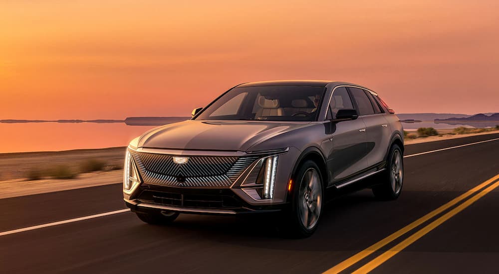 A silver 2022 Cadillac Lyriq EV is shown driving by the ocean during a sunset.