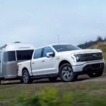 A white 2022 Ford F-150 Lightning is shown towing an airstream on an open road.
