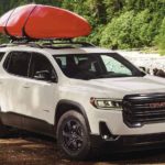 A white 2022 GMC Acadia is shown from the side parked next to a river with kayak on the roof.