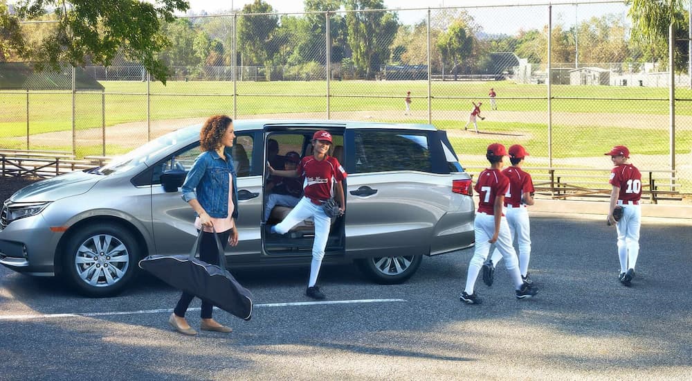 Children in baseball uniforms are getting out of a silver 2021 Honda Odyssey Elite next to a baseball field.