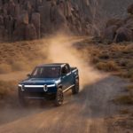 A blue Rivian R1T is shown driving on a dusty desert road.