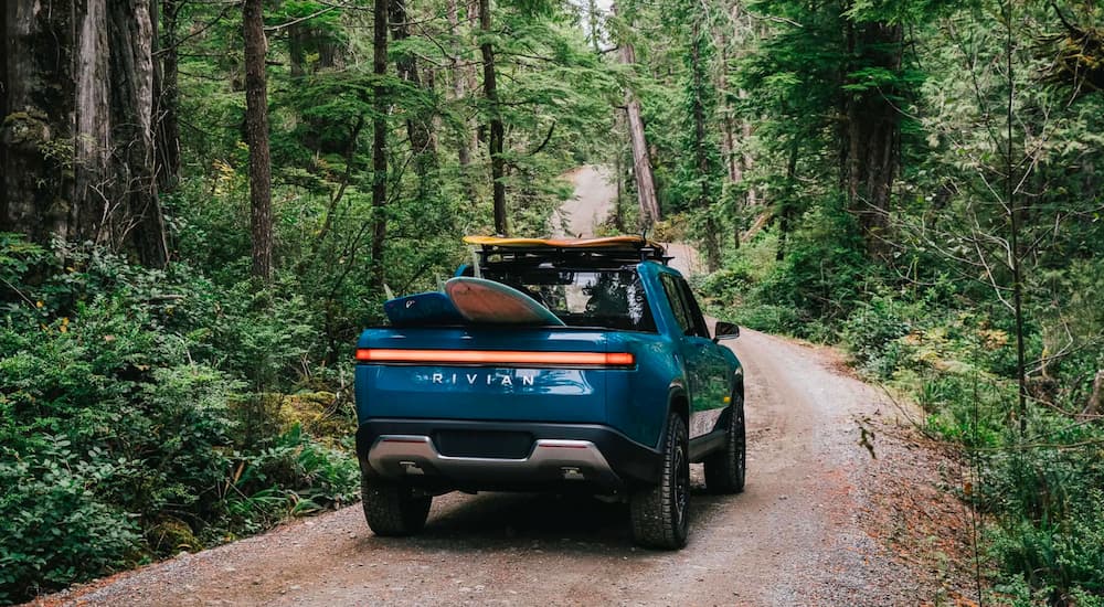 A blue Rivian R1T with surfboards in the bed driving is down a road in a forest.