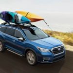 A blue 2022 Subaru Ascent with two kayaks on the roof rack is shown from a high angle driving by the ocean.