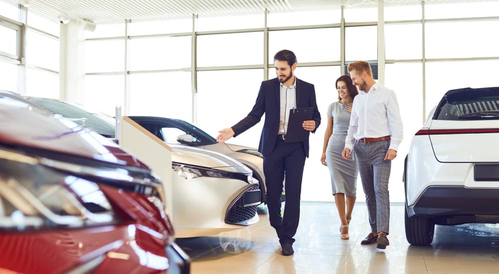 A couple and a salesman are shown looking at vehicles at a used car dealership.