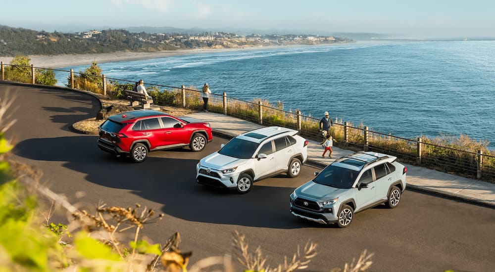 A red 2021 Toyota RAV4 Limited, a white XSE, and a blue Adventure Grade are shown from a high angle overlooking the ocean.