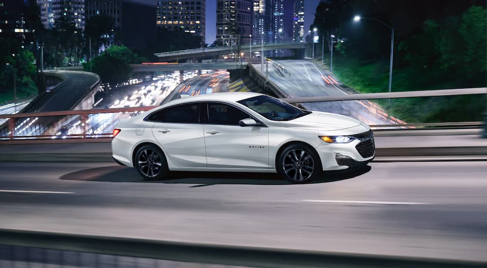 A white 2022 Chevy Malibu is shown driving on a city overpass at night.