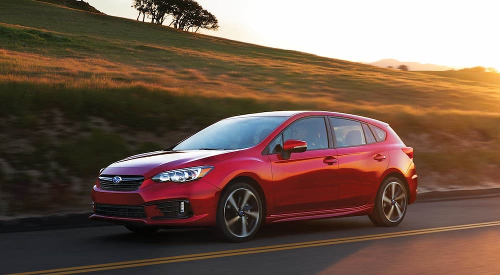 A red 2021 Subaru Impreza is shown driving on a road passing a grassy hill.