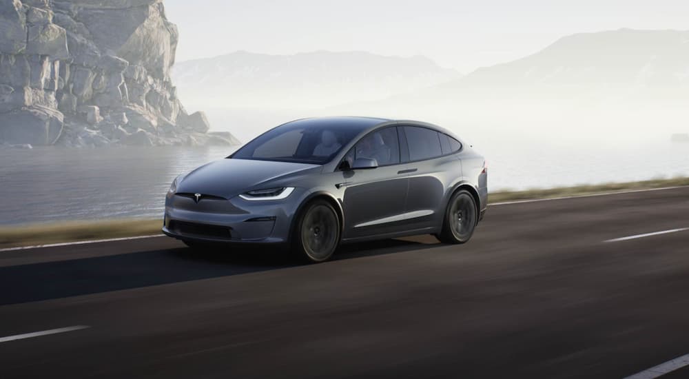 A grey 2021 Tesla Model X is shown driving on a highway near a body of water.