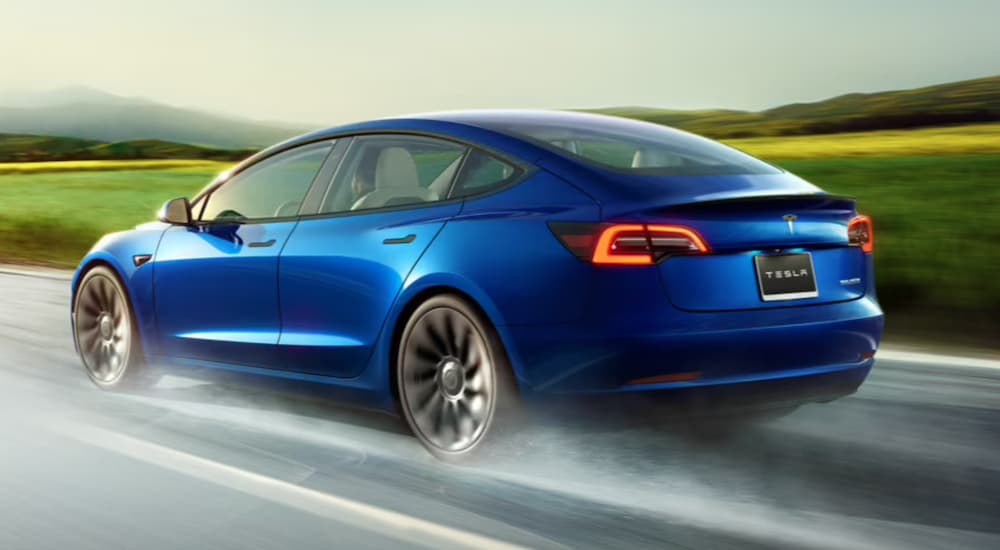 A blue Tesla Model 3 is shown from a rear angle driving on a highway.