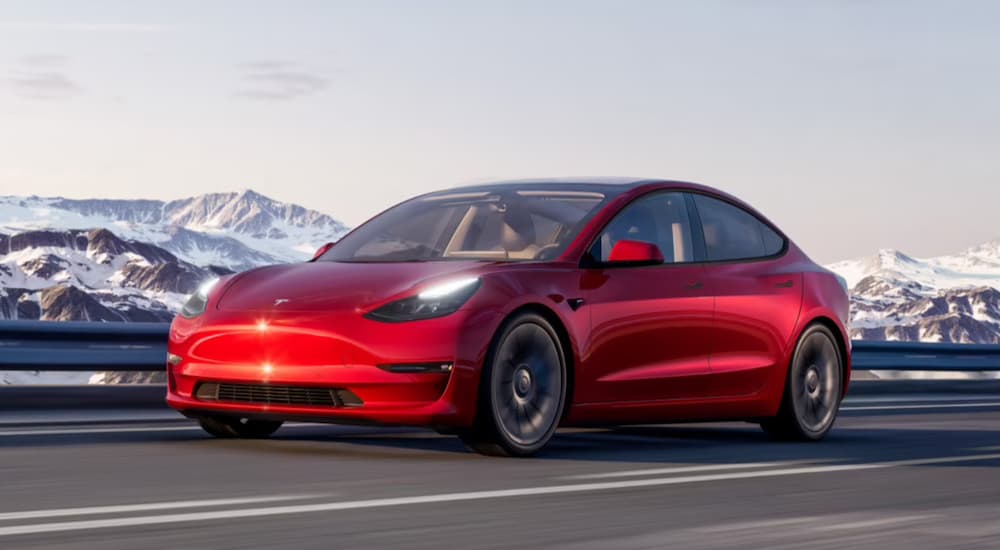 A red Tesla Model 3 is shown driving on a highway near mountains.