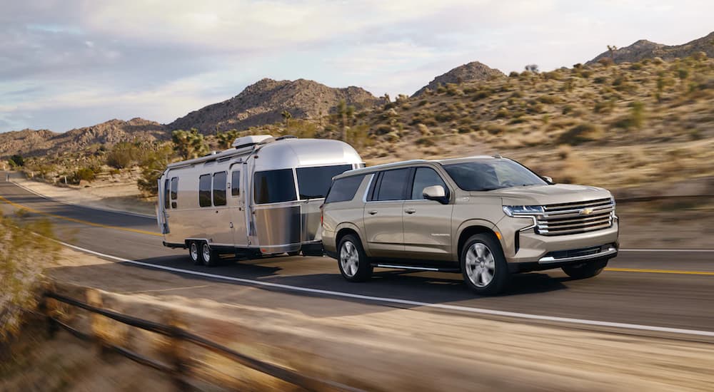 A tan 2022 Chevy Suburban is shown towing a camper on an open road in the desert.