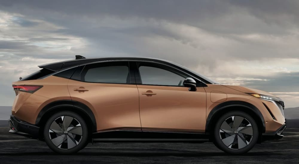 A gold 2023 Nissan Ariya is shown from the side parked under a cloudy sky.