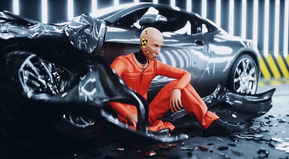 A crash test dummy in an orange jumpsuit is shown sitting against a crashed car.