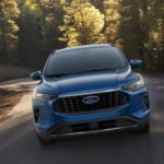 A blue 2023 Ford Escape is shown from the front driving on a winding road lined with trees.