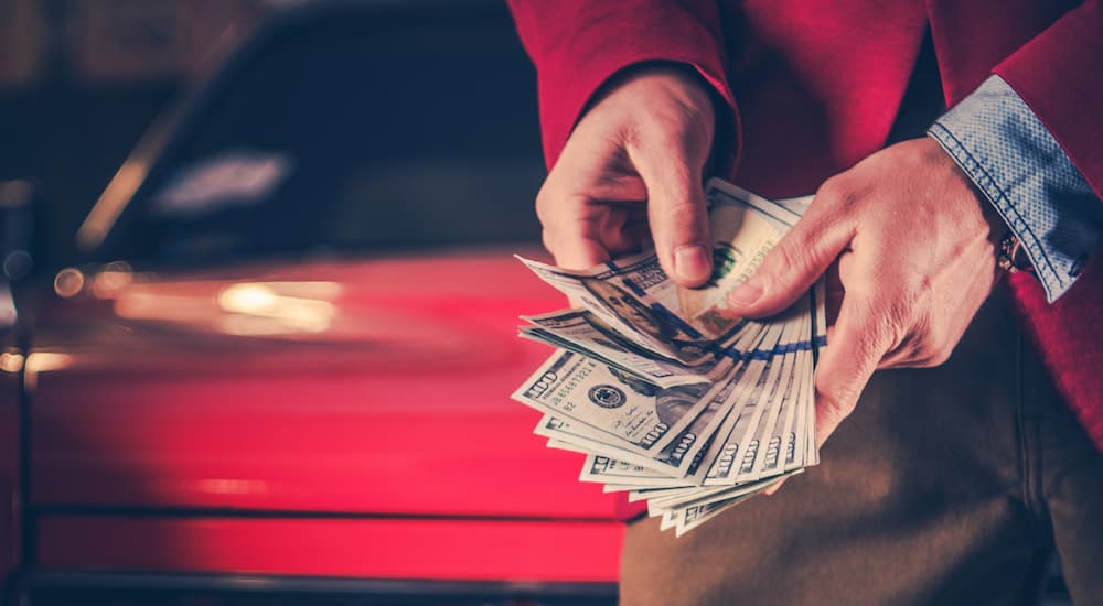 A person is shown holding a hand full of cash in front of a red car.