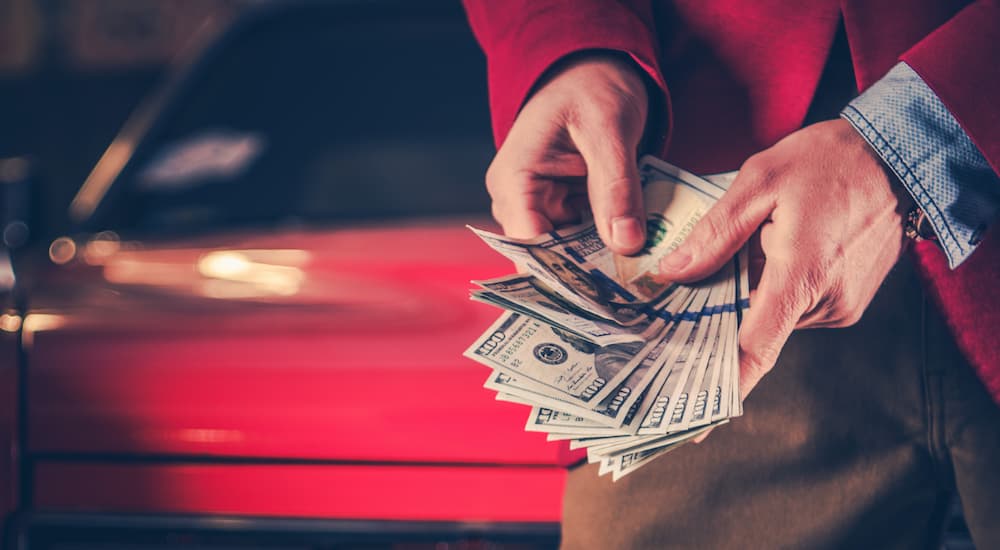 A person is shown holding a hand full of cash in front of a red car.