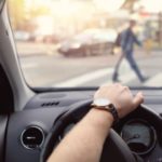 A person is shown with their hand on a steering wheel waiting for pedestrians to cross the street.