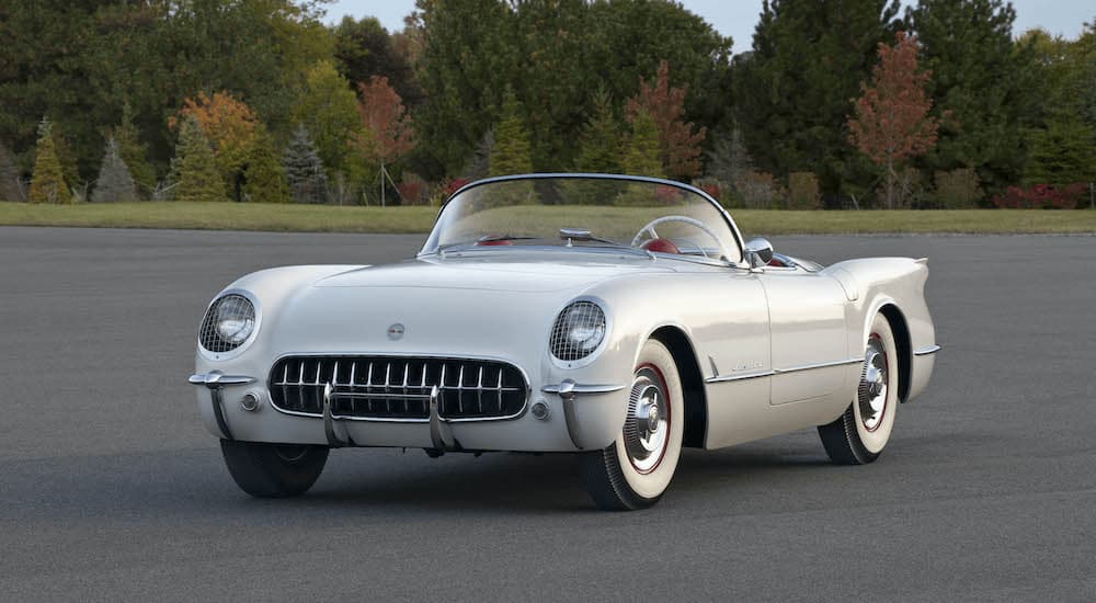 A white 1954 Chevy Corvette is shown parked in front of a line of trees.
