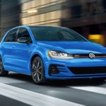 A blue 2021 Volkswagen Golf GTI is shown from the front driving through a city.