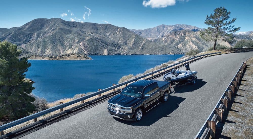 A black 2018 Chevy Silverado 1500 High Country is shown towing a boat next to a lake and mountains.