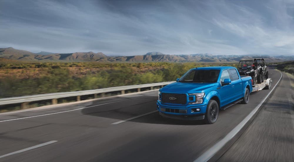 A blue 2019 Ford F-150 is shown towing ATVs on an empty road.