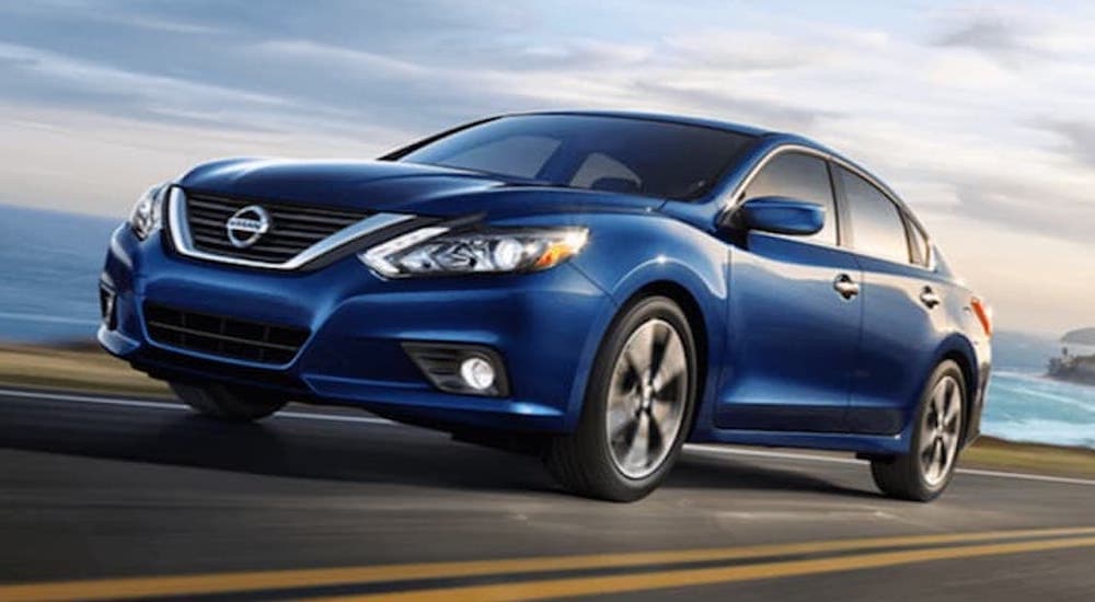 A blue 2018 Nissan Altima is shown from the side driving on an open road past a body of water.