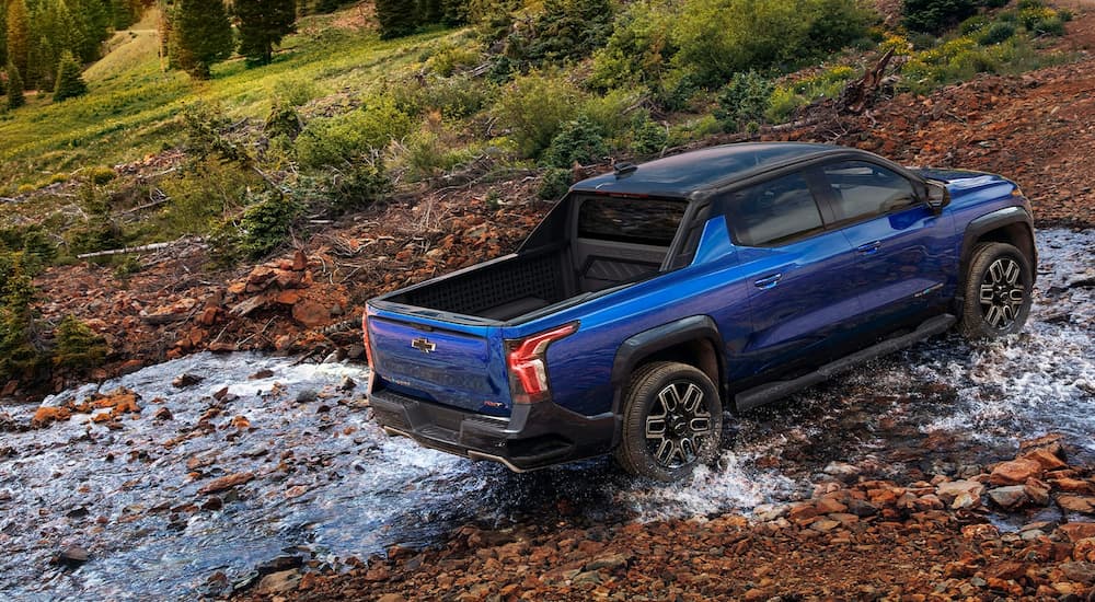 A blue 2024 Chevy Silverado EV is shown from the side driving through a small stream.