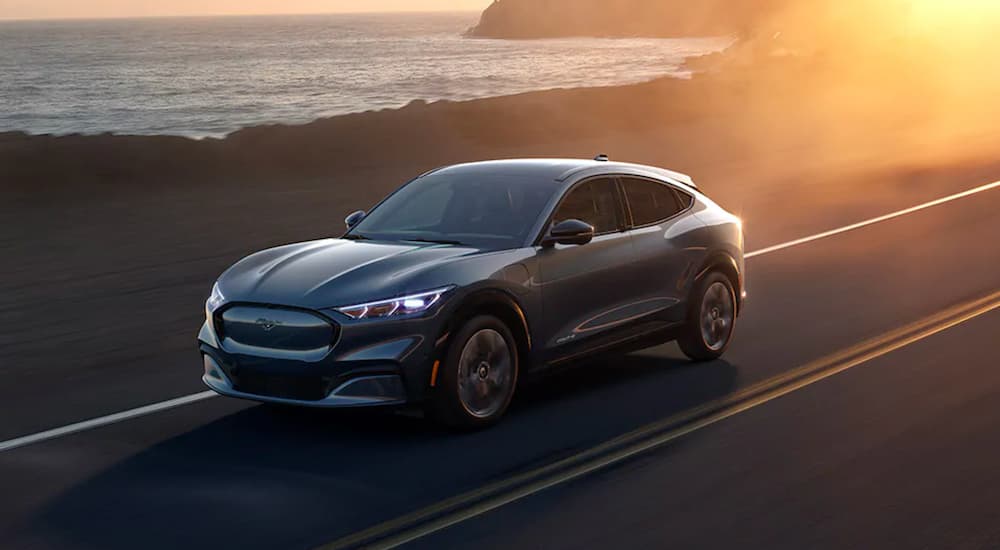 A grey 2021 Ford Mustang Mach-E is shown from the front at an angle driving on a coastal road at sunset.