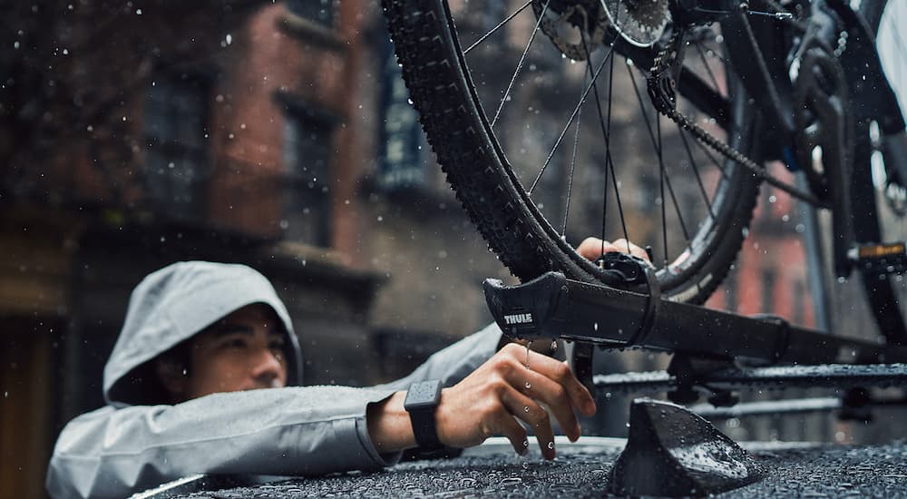 A person is shown securing a bike to the roof rack of a black 2022 Mazda CX-5 in the rain.