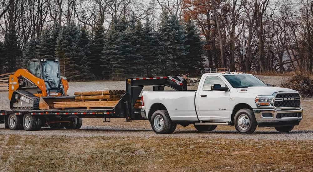 A white 2022 Ram 3500 is shown from the side while it tows a loader and some lumber.